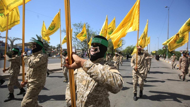 Masked individuals in camouflage uniforms march down a street carrying numerous yellow flags with green emblems.