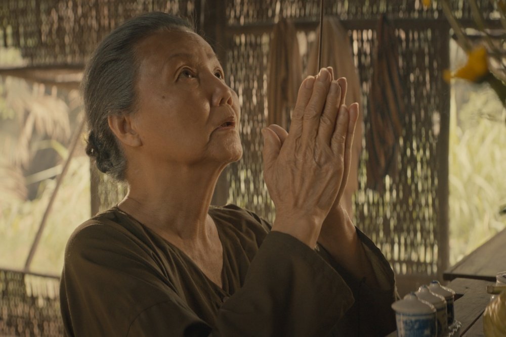 An elderly woman with gray hair stands with hands pressed together in a prayer-like gesture, gazing upward inside a bamboo structure with teacups on a nearby table.