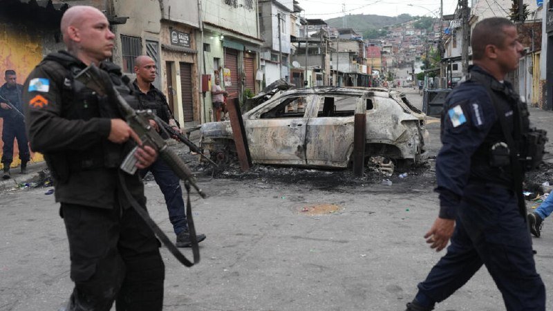 Armed officers patrol a street beside a burnt-out vehicle, with a hillside favela visible in the background.