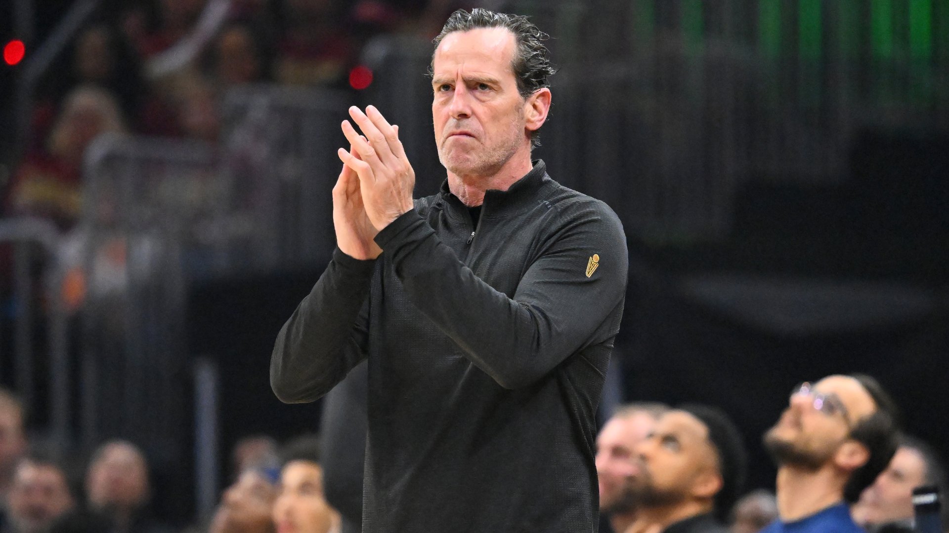 A gray-haired man in a black quarter-zip pullover claps his hands while standing courtside, with blurred spectators visible in the background.