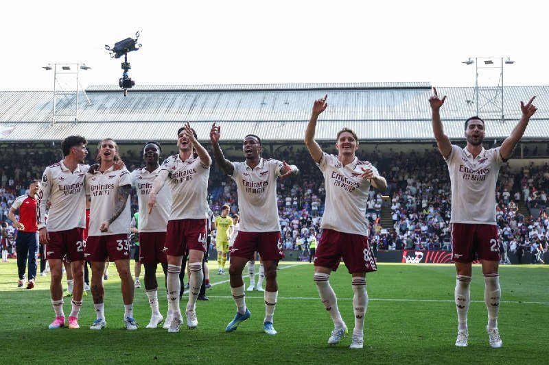 Arsenal players in white and maroon kits celebrate with arms raised toward fans in a stadium stand after a match.
