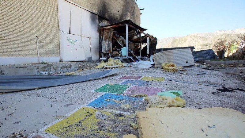 A burned building shows blackened walls and a collapsed roof, with debris, insulation, and a colorful painted floor scattered across the ground in front.