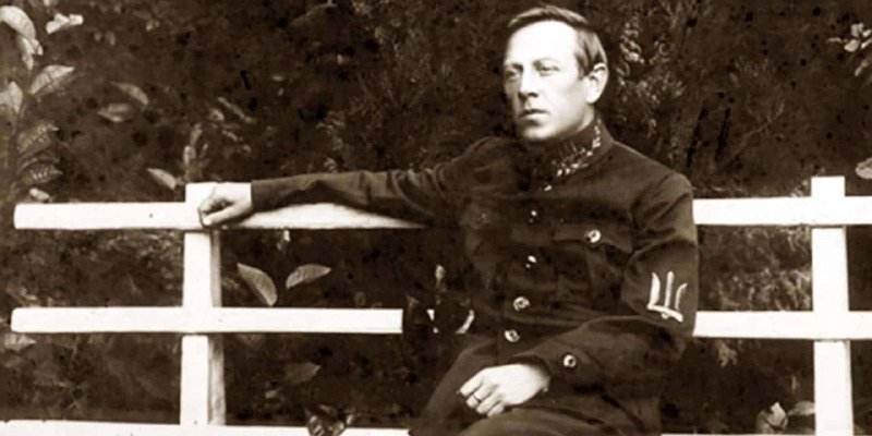Sepia photograph showing a man in a dark uniform seated beside a white wooden fence, with foliage visible in the background.