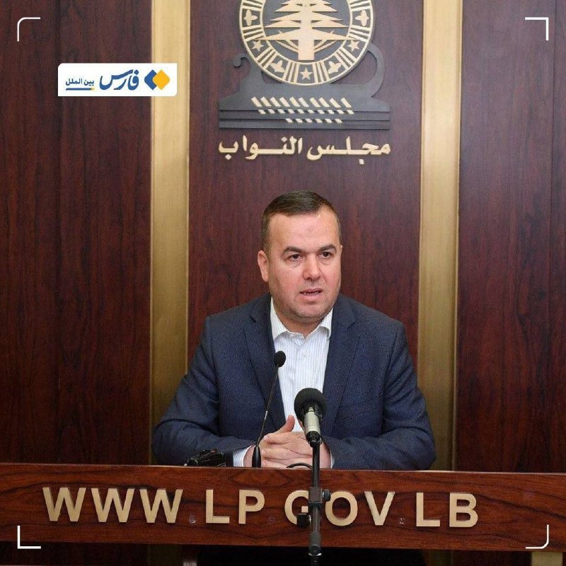 A man in a blue suit speaks at a wooden podium bearing "WWW.LP.GOV.LB," with an emblem and Arabic text mounted on the wood-paneled wall behind him.