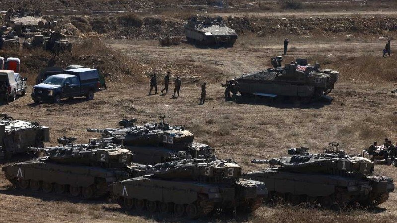 Military personnel stand among multiple armored tanks and vehicles positioned in a dry, grassy field.