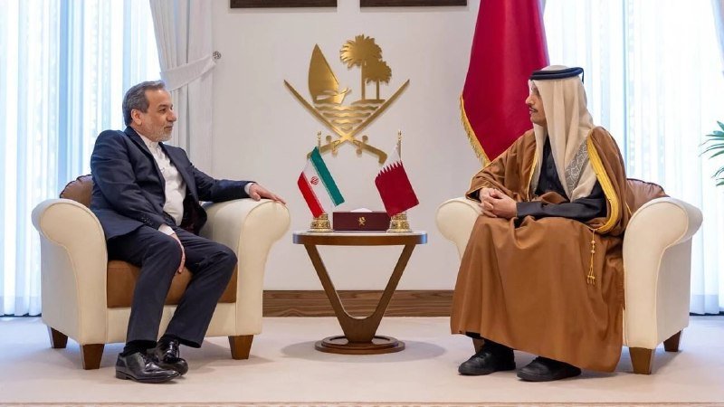 Two men in formal attire sit facing each other in armchairs across a small table displaying Iranian and Qatari flags, with an emblem on the wall behind.