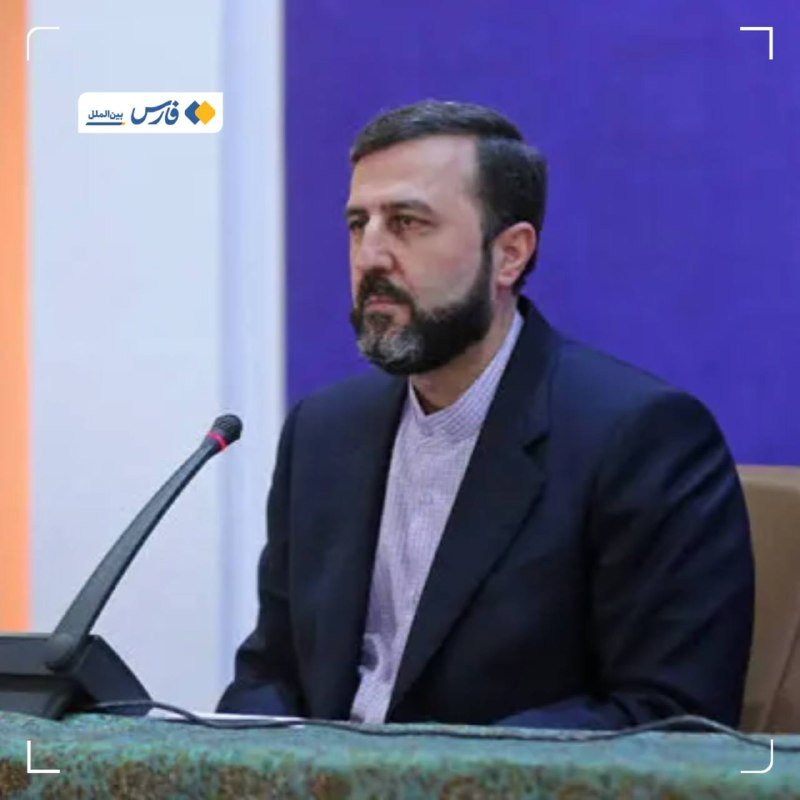 A bearded man in a dark blazer and light shirt sits at a desk with a microphone, speaking against a blue backdrop with a logo visible in the corner.