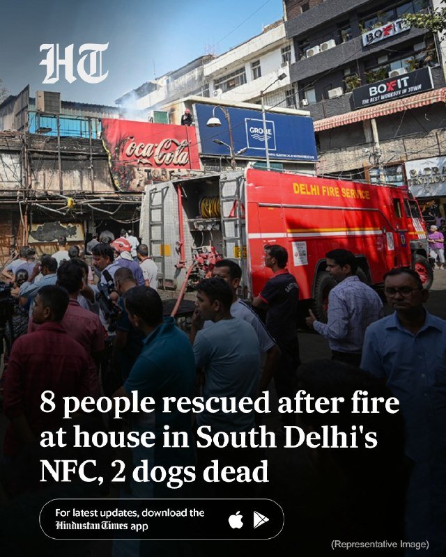 Red Delhi Fire Service truck parked on a crowded street as onlookers gather near a building with a Coca-Cola billboard, smoke visible in the background.
