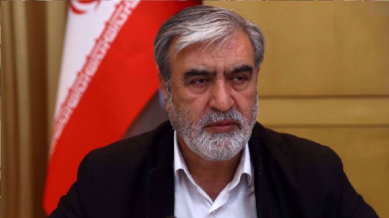 A gray-haired, bearded man in a dark suit jacket and white shirt sits in front of a red and white flag.