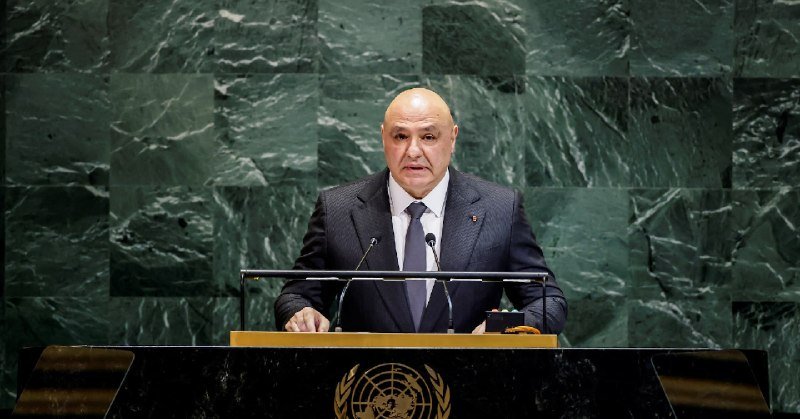 A bald man in a dark suit and blue tie stands at a podium bearing the UN emblem, speaking before a green marble backdrop.