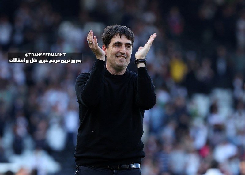 A smiling man in a black sweater claps his hands while standing before a blurred crowd of spectators in stadium seating.