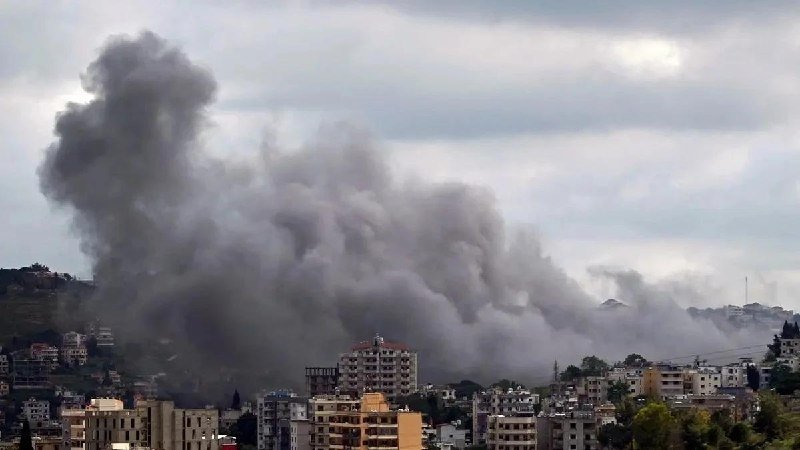 Thick plumes of gray smoke rise above a densely built hillside city under a partly cloudy sky.