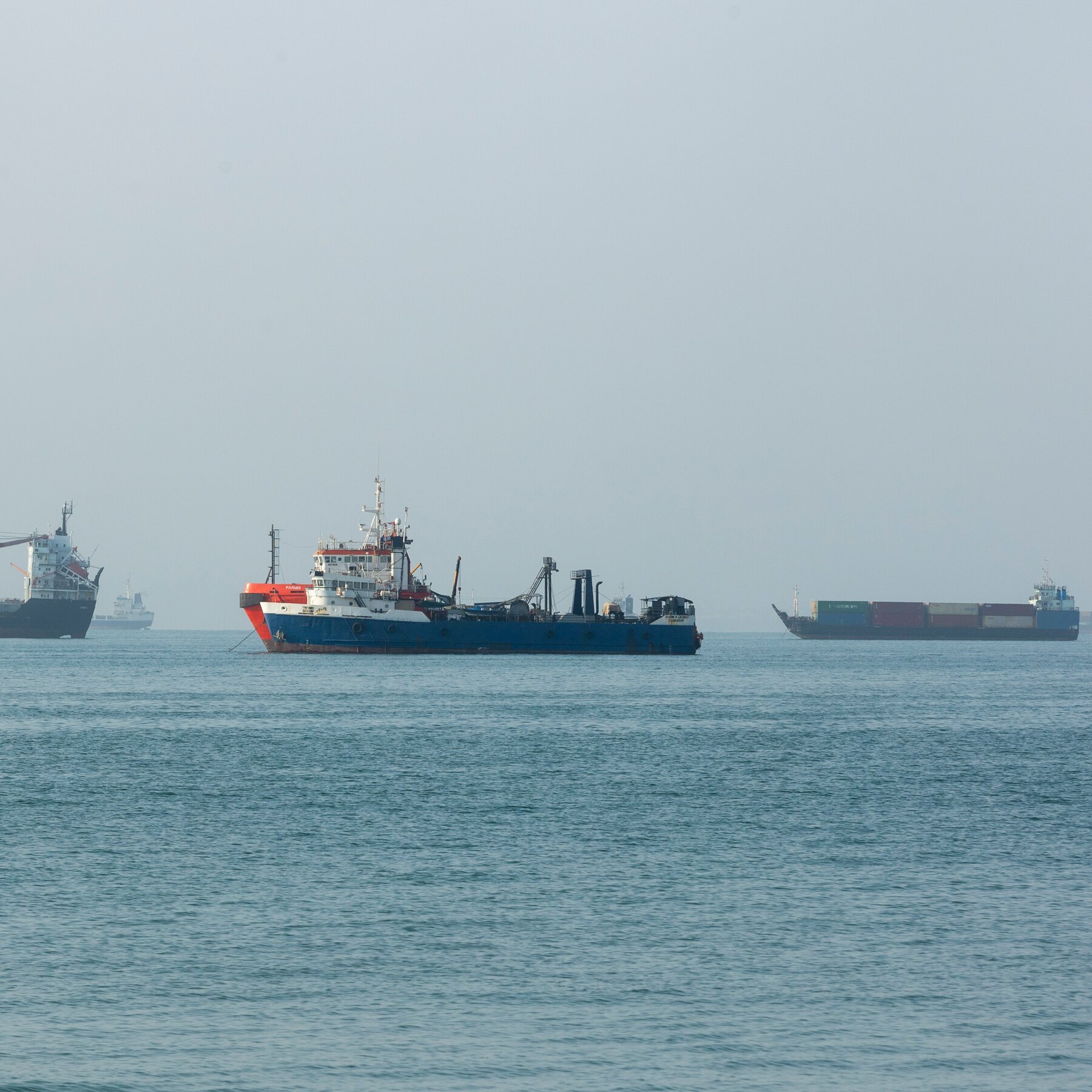A blue and red industrial vessel sits anchored in calm gray-blue waters, with a container ship visible to the right and another vessel partially seen on the left horizon.