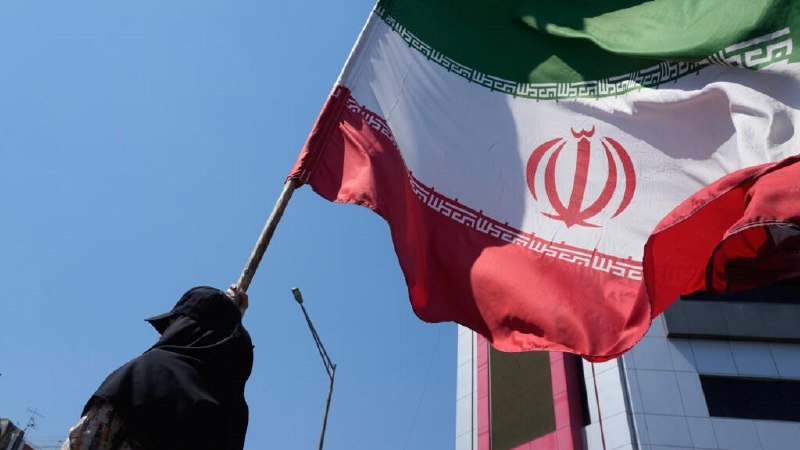 A person in dark clothing waves a large Iranian flag against a clear blue sky, with a building visible in the background.