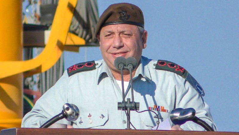 A military officer in a light blue uniform with insignia and a beret stands at a podium equipped with multiple microphones, appearing to address an audience outdoors.