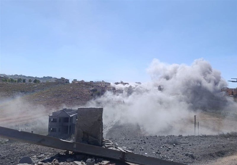 A large plume of dust rises over a hillside with scattered buildings, surrounding a partially collapsed structure amid rocky, debris-covered terrain under a clear blue sky.
