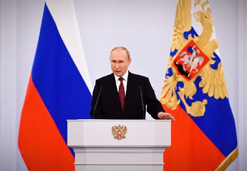 A man in a dark suit and red tie speaks at a podium bearing a double-headed eagle emblem, flanked by the Russian flag and a presidential standard.