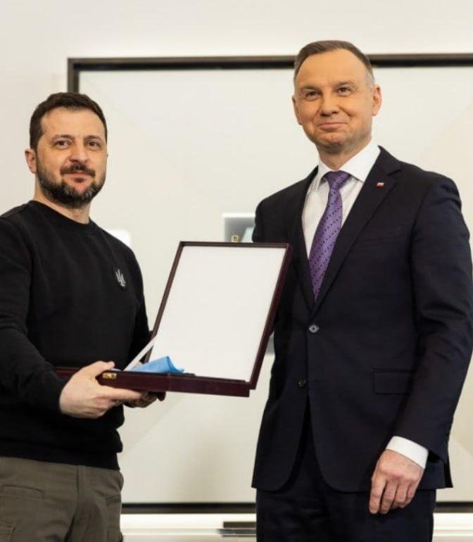 Two men stand indoors, one in a dark sweater holding open a presentation box while the other, in a dark suit and purple tie, looks on.