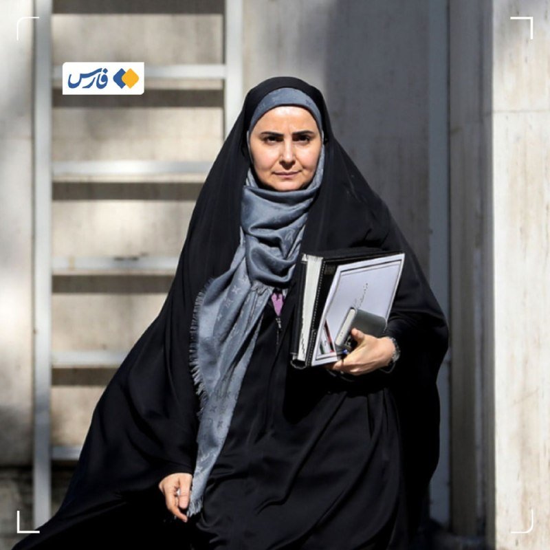 A woman wearing a black chador and gray headscarf walks outdoors carrying binders and a mobile phone, with a metal ladder visible behind her.