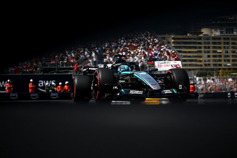 A black and teal Formula 1 race car with "Petronas Syntium" branding drives on a dark track, with crowded grandstands and trackside marshals visible in the background.