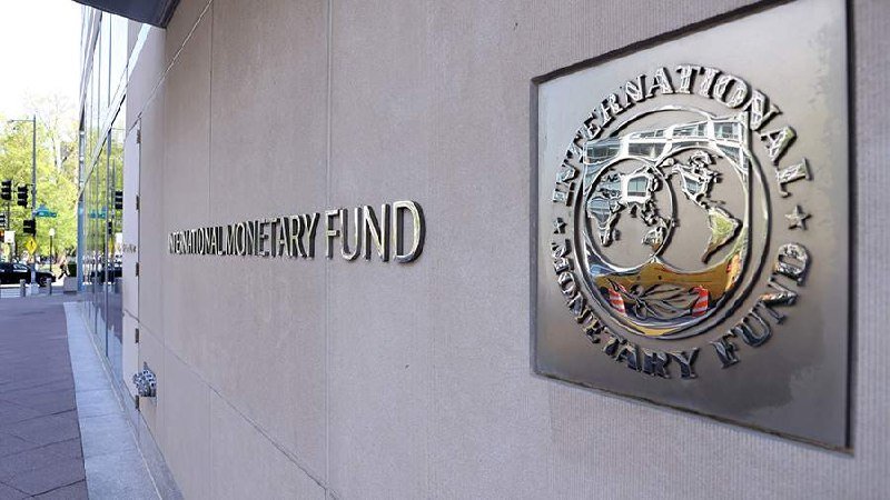 A metallic plaque and engraved lettering display the "International Monetary Fund" name and logo on a building's exterior stone wall along a city sidewalk.