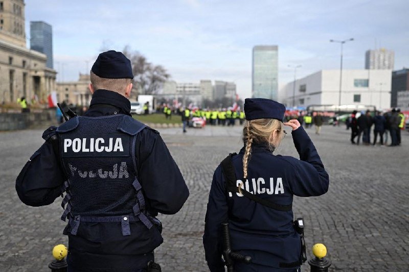 Two Polish police officers in "POLICJA" vests stand with their backs to the camera, observing a crowd and a line of additional officers in a city square.