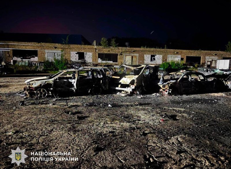 Burned-out vehicles sit in a debris-strewn lot in front of a damaged brick building at night, with a "Національна поліція України" logo visible.