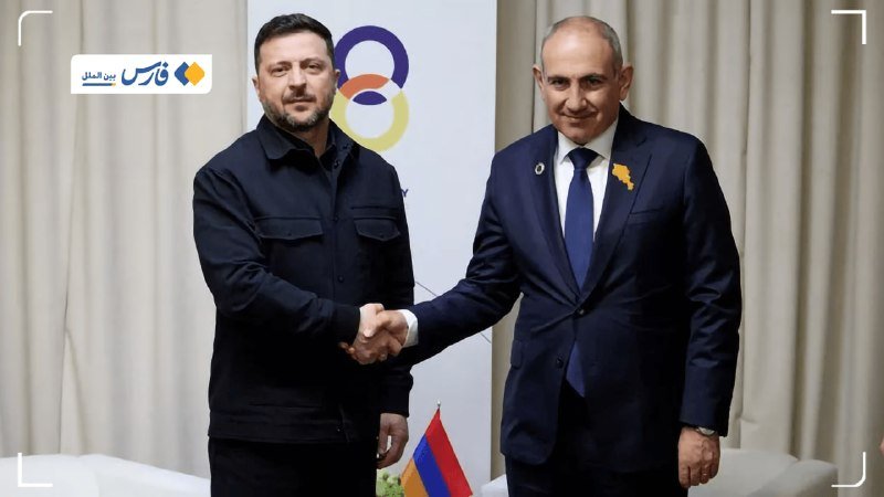 Two men in dark suits shake hands indoors in front of light curtains, with a small red, blue, and orange tricolor flag visible below.