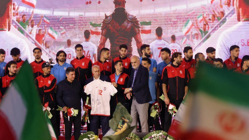 A group of men in red and white tracksuits gather on a stage holding a jersey marked "12," with bouquets and Iranian flags in the foreground and a stadium backdrop.