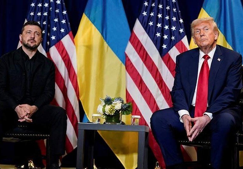 Two men sit in chairs in front of alternating Ukrainian and American flags, with a small floral arrangement on a table between them.