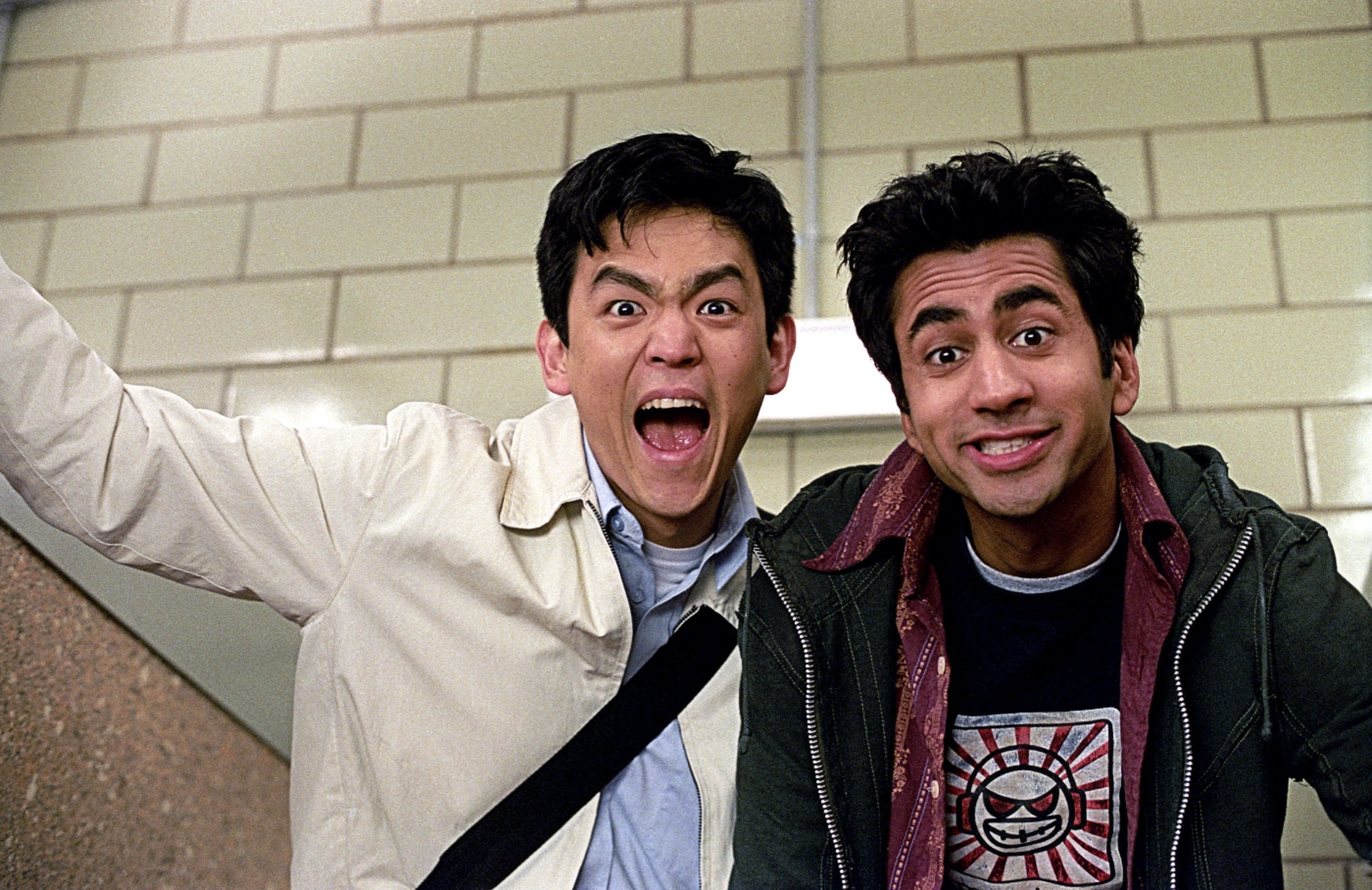 Two men pose for the camera indoors against a beige brick wall, one with an open-mouthed expression and the other smiling, wearing a graphic t-shirt.
