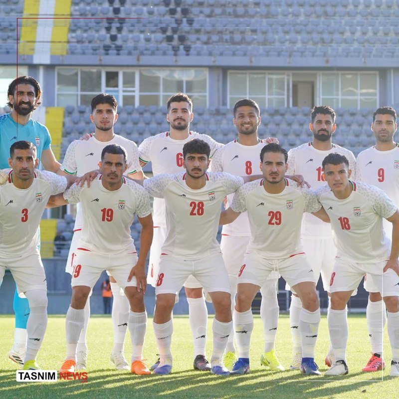 Eleven soccer players in white and teal uniforms pose together for a team photo on a grass field, with empty stadium seating visible behind them.