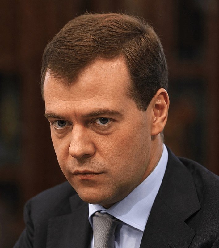 A man with short brown hair, wearing a dark suit, light blue shirt, and gray tie, looks to the side with a serious expression against a blurred wooden background.