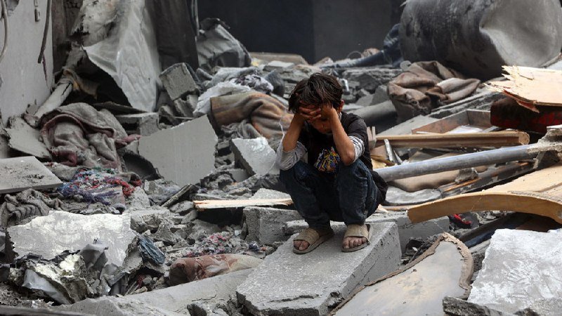 A young boy squats on a concrete slab amid extensive rubble, covering his face with both hands amid the debris of a destroyed building.