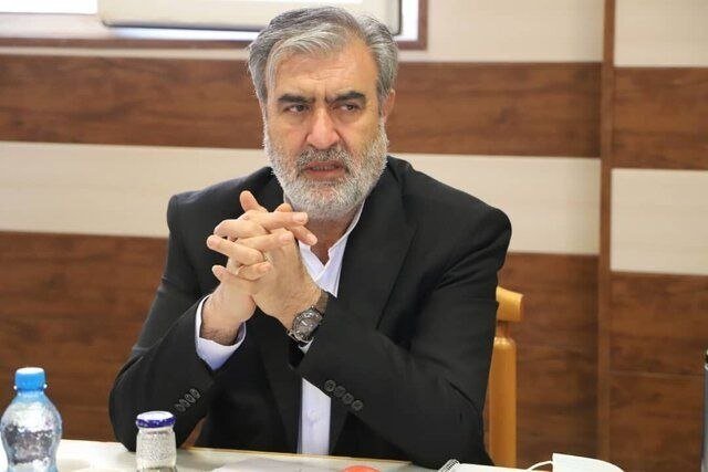 A gray-bearded man in a dark suit sits at a table with hands clasped, a water bottle and glass beside him, against a wood-paneled wall.