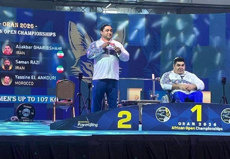 Two men in white and blue tracksuits stand on numbered podiums (2 and 1) at the Oran 2026 African Open Championships powerlifting stage, with a screen listing Iranian and Moroccan competitors behind them.