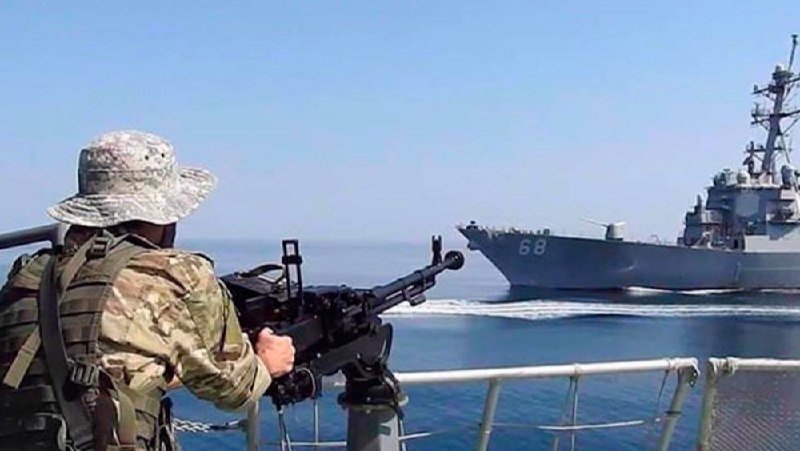 A soldier in camouflage and a boonie hat mans a mounted machine gun aboard a vessel, facing a naval warship marked "68" on the open sea.