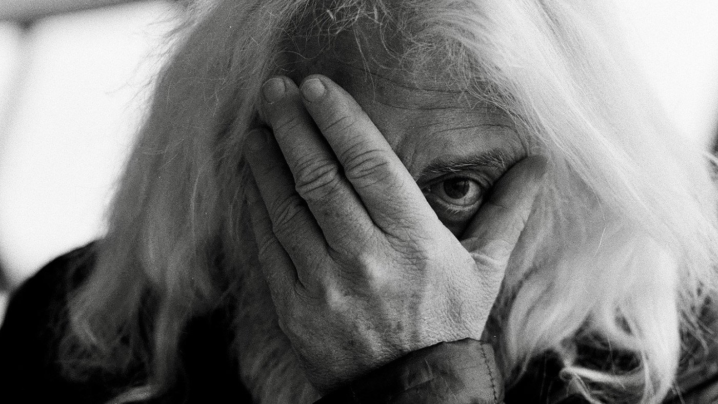 A black-and-white close-up shows a person with long, light hair covering most of their face with one hand, revealing only one eye.