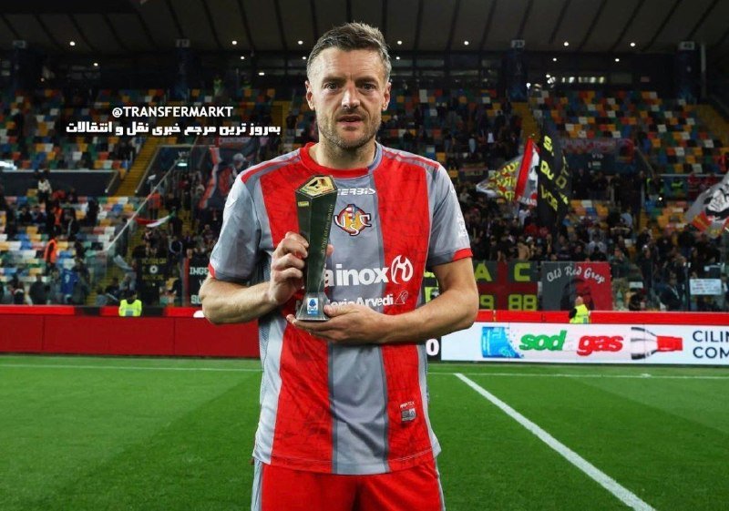 A bearded man in a red and grey striped jersey holds a small trophy on a soccer pitch, with stadium crowds visible behind him.