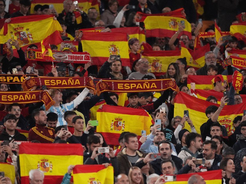 Crowd of spectators in stadium stands wave red and yellow Spanish flags and hold up "ESPAÑA" scarves while taking photos with their phones.