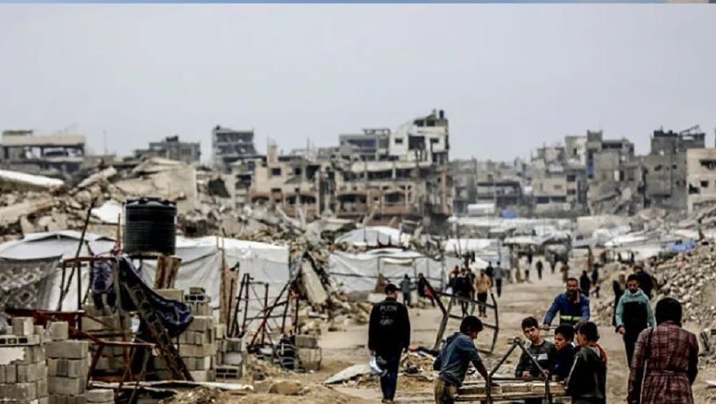 People walk through a landscape of heavily damaged buildings and rubble, with makeshift tents and construction materials scattered among the debris.