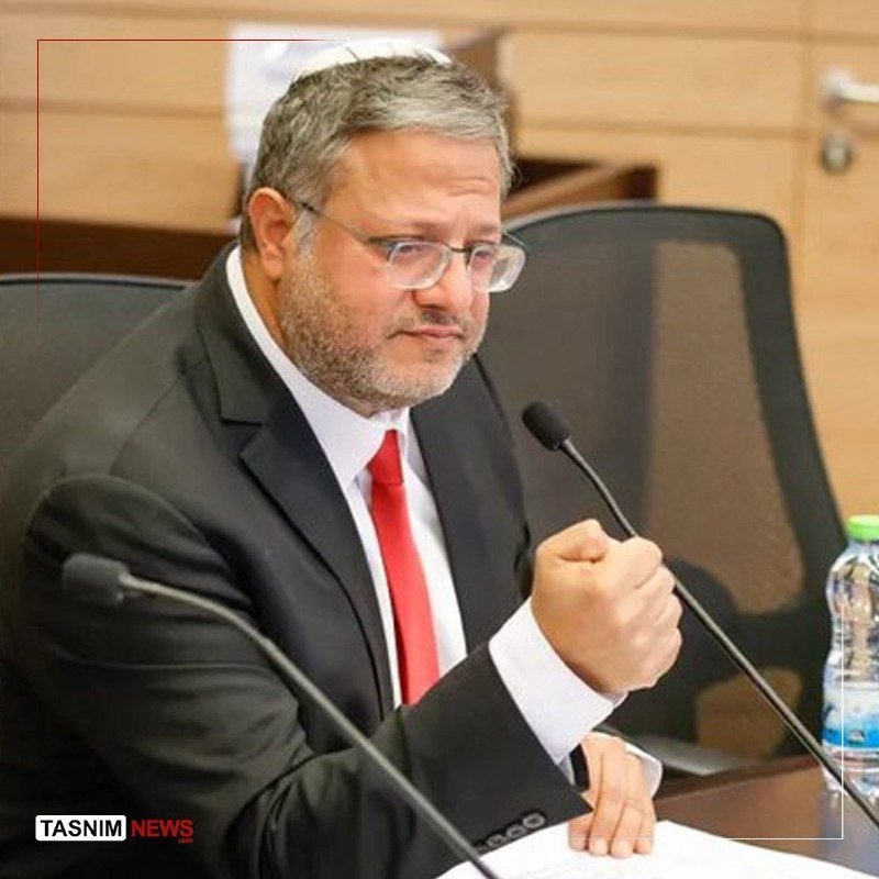 A bespectacled, bearded man in a black suit and red tie sits at a desk with microphones, raising one clenched fist, with a "Tasnim News" logo visible.