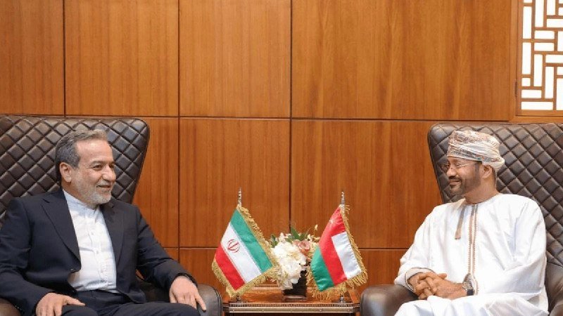 Two men in traditional and formal attire sit smiling in leather chairs across from each other, with the Iranian and Omani flags displayed on a table between them against a wood-paneled wall.
