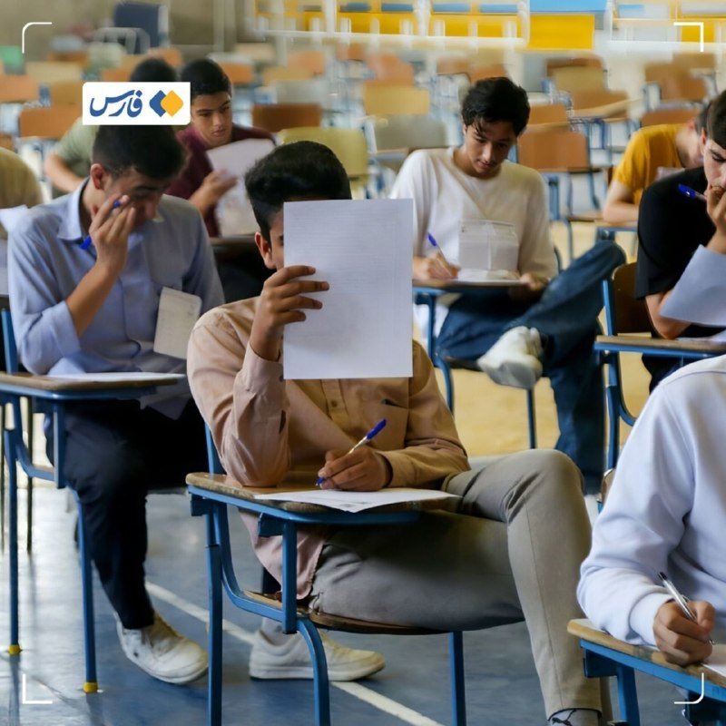 Students seated at individual desks in a large hall, writing on papers and holding up sheets while taking an examination.