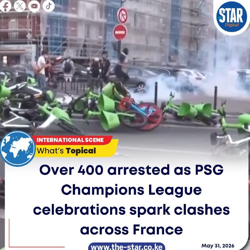 Smoke rises over a barricade of toppled green rental bikes on a city street, with uniformed officers and bystanders visible in the background.