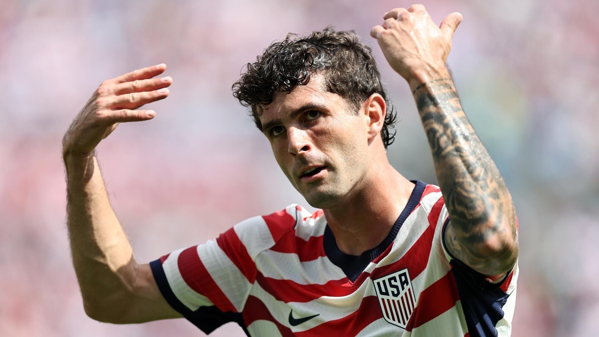 A tattooed soccer player in a red, white, and blue striped USA jersey raises both arms while looking forward against a blurred stadium background.