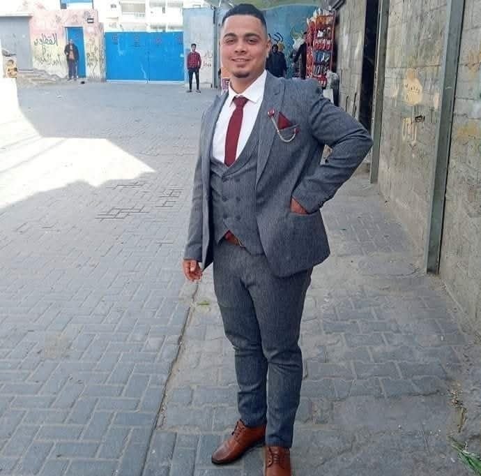 A man in a gray three-piece suit with a red tie and pocket square stands smiling on a paved street, with children and shop displays visible in the background.