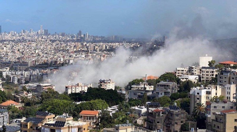 Plumes of smoke rise above densely packed residential buildings in a sprawling city.