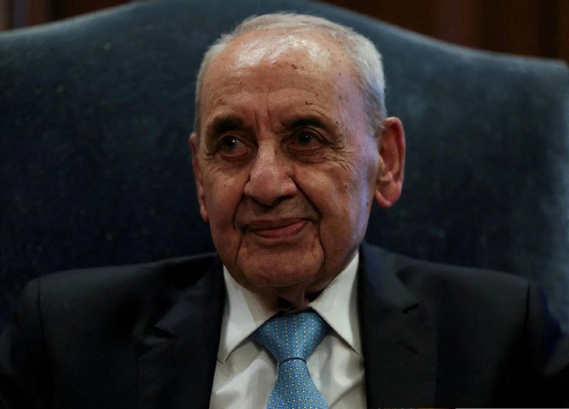 An elderly man with gray hair, wearing a dark suit and light blue tie, sits in a high-backed chair with a slight smile.
