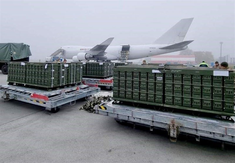 Pallets of green military crates sit on flatbed dollies on an airport tarmac, with a large gray military aircraft parked in the foggy background.